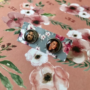 May include: A pair of silver stud earrings with a round glass cabochon featuring a printed image of a man and a woman. The earrings are on a blue and white floral background.