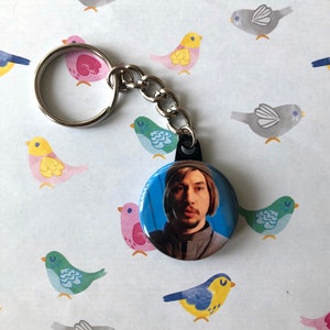 May include: A silver keychain with a round button featuring a photo of Adam Driver. The button is black with a silver rim. The background of the image is a blue wall.