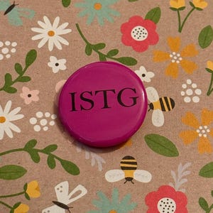 May include: A magenta button with the letters "ISTG" in black. The button is set against a floral background with flowers, bees, and butterflies in various colors.