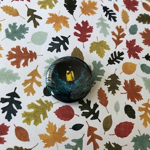 May include: A round button pin with a dark blue background and a yellow figure in a yellow raincoat standing in the middle of a dark forest. The figure is holding a small yellow umbrella.