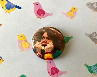 May include: A round button with a picture of a woman with brown hair and a red and yellow jacket, looking scared and holding her hands to her face. The background is blurry.