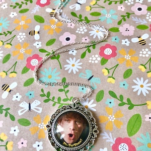 May include: A silver necklace with a round pendant featuring a photo of a person with a surprised expression. The pendant has a decorative silver frame and the text "Po-Ta-Toes!" printed in yellow.