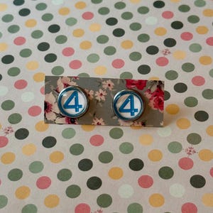 May include: A pair of round stud earrings featuring the number 4 in blue against a white background, set in a silver-colored metal. The earrings are displayed on a floral patterned card, set against a polka dot background.