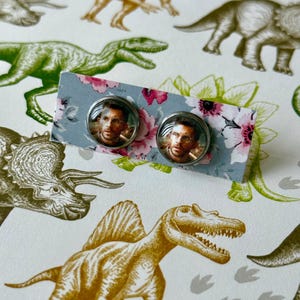 May include: A pair of round stud earrings featuring a portrait of a person. The earrings are set on a floral patterned card. The background is a white surface with dinosaur illustrations in green and gold.