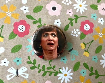 May include: A round button with a picture of a woman with brown hair and a surprised expression. The background is a floral pattern with pink, yellow, and white flowers.