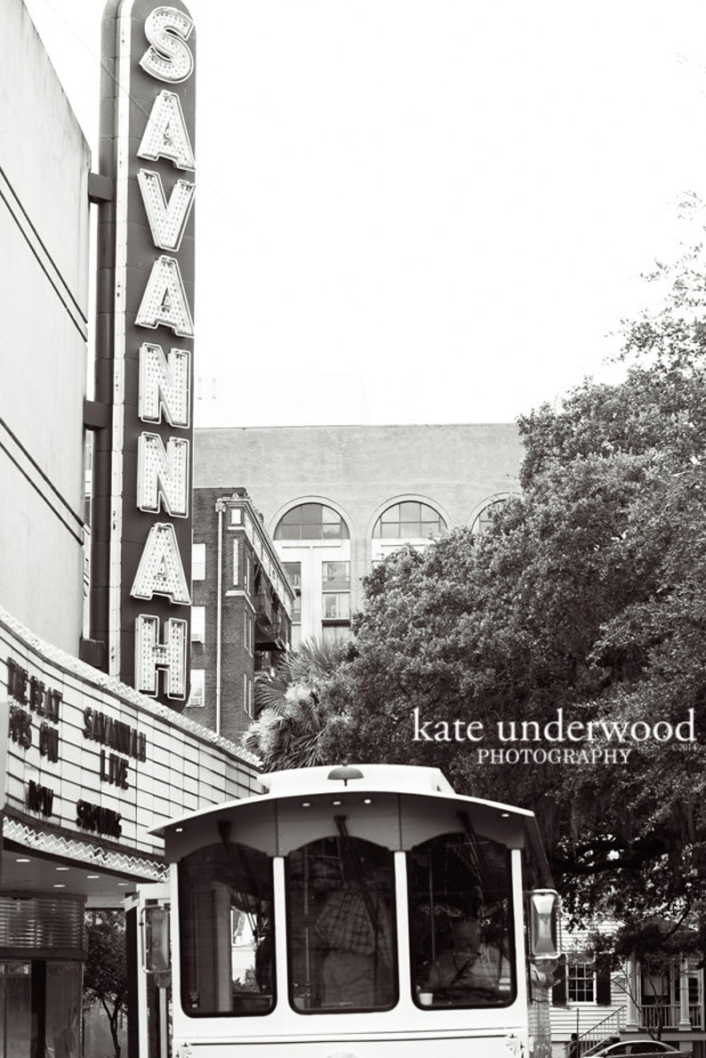 May include: A black and white photo of a vintage trolley car in front of a building with a sign that reads "SAVANNAH". The trolley car is empty and has a large window in the front. The building is old and has a brick facade. The photo is taken from a low angle, looking up at the trolley car.