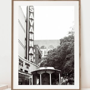 May include: Black and white photograph of a vintage movie theater marquee in Savannah, Georgia. The marquee reads "SAVANNAH" in large, illuminated letters. A trolley car is parked in front of the theater.