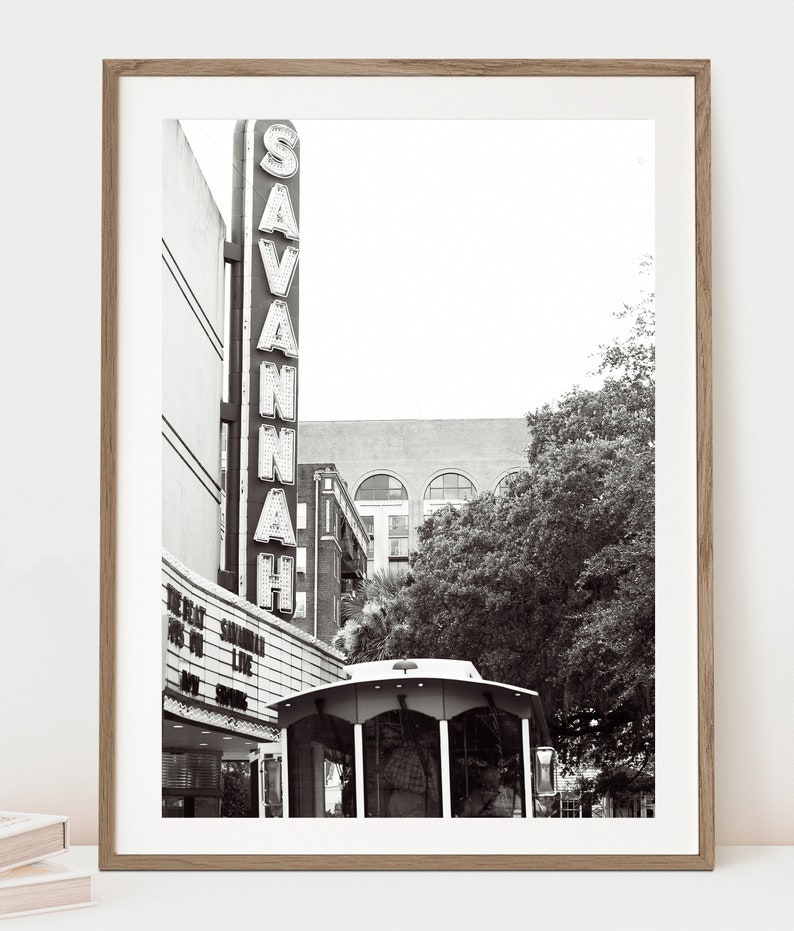 May include: Black and white photograph of a vintage movie theater marquee in Savannah, Georgia. The marquee reads "SAVANNAH" in large, illuminated letters. A trolley car is parked in front of the theater.
