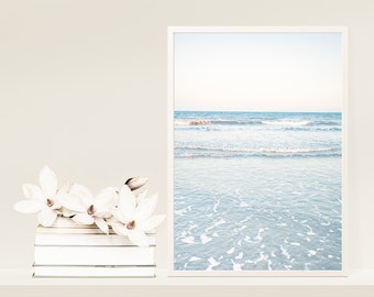 May include: A framed photograph of a beach scene with white sand and blue water with waves crashing on the shore. The photo is on a shelf with a stack of white books and a cluster of white magnolia flowers.