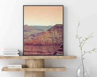 May include: Framed print of the Grand Canyon. The landscape features layers of red and brown rock formations under a peach-colored sky. The print is displayed on a wooden shelf with books and a glass vase with greenery.