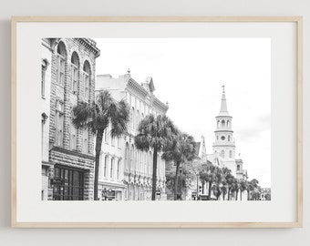 Op de afbeelding: Een zwart-witfoto van een straatbeeld in Charleston, South Carolina. De afbeelding toont een rij historische gebouwen met palmbomen langs de straat. Een hoge kerktoren is zichtbaar op de achtergrond.