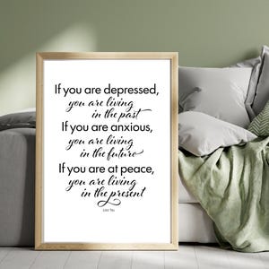May include: A framed print with a light wood frame. The print features black cursive text on a white background with a quote by Lao Tzu about living in the present. The frame is rectangular and the print is displayed on a bed with pillows and a green blanket.