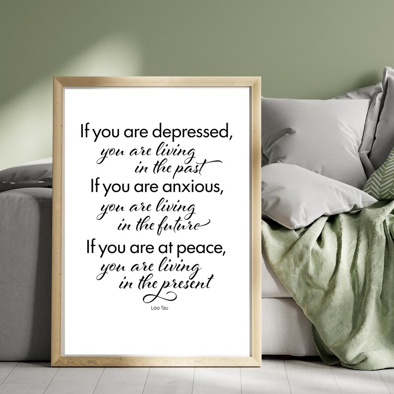 May include: A framed print with a light wood frame. The print features black cursive text on a white background with a quote by Lao Tzu about living in the present. The frame is rectangular and the print is displayed on a bed with pillows and a green blanket.