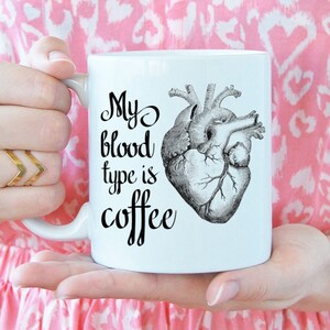 May include: White coffee mug with a black and white anatomical heart illustration and the text "My blood type is coffee".