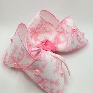 May include: A large, decorative hair bow in white fabric with a pink floral pattern and pink trim. The bow is embellished with pink beads and a small pink fabric flower in the center.