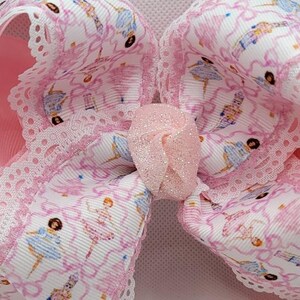 Puede incluir: Un lazo grande y rosa para el cabello con estampado de bailarina. El lazo presenta una cinta blanca con un patr&oacute;n de bailarinas en rosa y azul, acentuada con un ribete de encaje rosa y un centro brillante.