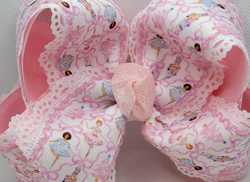 Puede incluir: Un lazo grande y rosa para el cabello con estampado de bailarina. El lazo presenta una cinta blanca con un patr&oacute;n de bailarinas en rosa y azul, acentuada con un ribete de encaje rosa y un centro brillante.