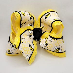 May include: A large hair bow featuring a cream-colored fabric with a repeating pattern of yellow and black bees and yellow honeycomb outlines. The edges are trimmed with yellow lace and black piping, and the center is cinched with a black ribbon knot.