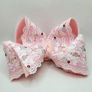 Puede incluir: Un lazo grande para el pelo, de color rosa, con un estampado de bailarina en blanco y rosa. El lazo tiene un ribete de encaje y una cinta de raso en el centro. El lazo mide aproximadamente 15 cm de ancho.