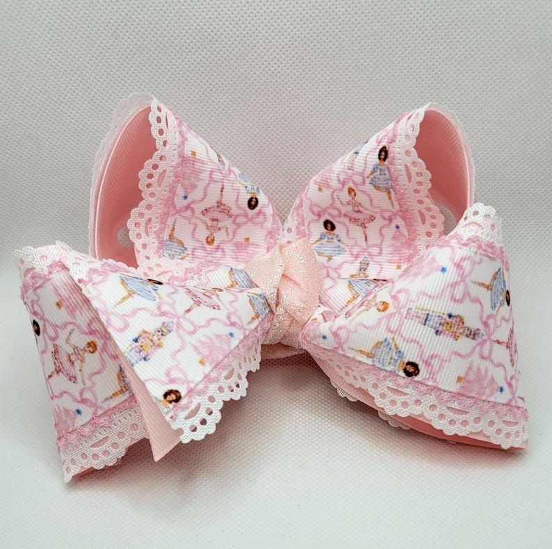 Puede incluir: Un lazo grande para el pelo, de color rosa, con un estampado de bailarina en blanco y rosa. El lazo tiene un ribete de encaje y una cinta de raso en el centro. El lazo mide aproximadamente 15 cm de ancho.