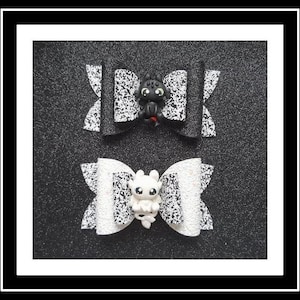 May include: Two decorative hair bows. One is black with a white speckled pattern and a black dragon figurine. The other is white with a white speckled pattern and a white dragon figurine. The bows are on a black background.