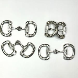 May include: Five silver-toned metal buckles with clear rhinestone embellishments. The buckles are in various shapes and sizes, including two pairs of matching buckles.