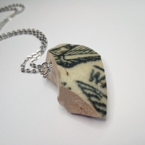 May include: A silver chain necklace with a pendant made from a broken piece of ceramic with black and white markings. The ceramic piece has a rough, uneven texture.