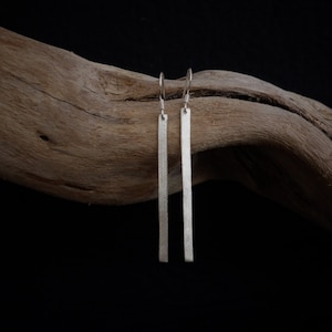 May include: A pair of silver bar earrings with a textured finish. The earrings hang vertically, suspended from small hooks. The background is a dark, neutral tone, with a piece of driftwood.