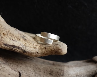 May include: A silver ring with a hammered finish, twisted into a unique design. The ring is displayed on a piece of driftwood.