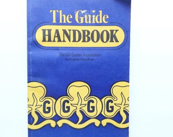 May include: A vintage blue handbook titled "The Guide Handbook" with gold lettering and design elements. The cover features the text "The Girl Guides Association" and the address "Buckingham Palace Road".