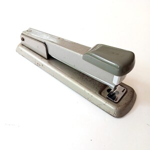 May include: A silver and gray metal stapler with a worn finish. The stapler has a lever on the top and a base with a metal plate.