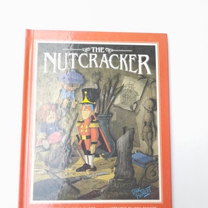 May include: A red, orange, and yellow book cover with the title "The Nutcracker" in black text. The cover features an illustration of a toy soldier with a red and gold uniform, holding a sword. The book is by Ronald Kidd and illustrated by Rick Reinert.
