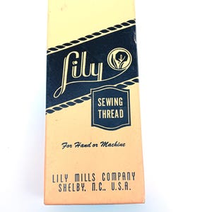 May include: A vintage box of Lily Sewing Thread. The box is rectangular with a cream and navy blue colour scheme. The word "Lily" is in a cursive font, and the text "Sewing Thread" is in a rectangular box. The text "For Hand or Machine" is also visible.