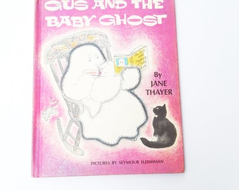 May include: A pink book cover with the title "Gus and the Baby Ghost" in white text. The cover features a cartoon illustration of a ghost sitting in a rocking chair, reading a book. A black cat sits on the floor to the right of the ghost. The text "By Jane Thayer" and "Pictures by Seymour Fleishman" are printed below the illustration.