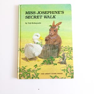May include: A children's book titled "Miss Josephine's Secret Walk" by Yuji Kobayashi. The cover features a duck, a rabbit, and a mouse near a woven basket. The background is a yellow-green field with pink flowers.
