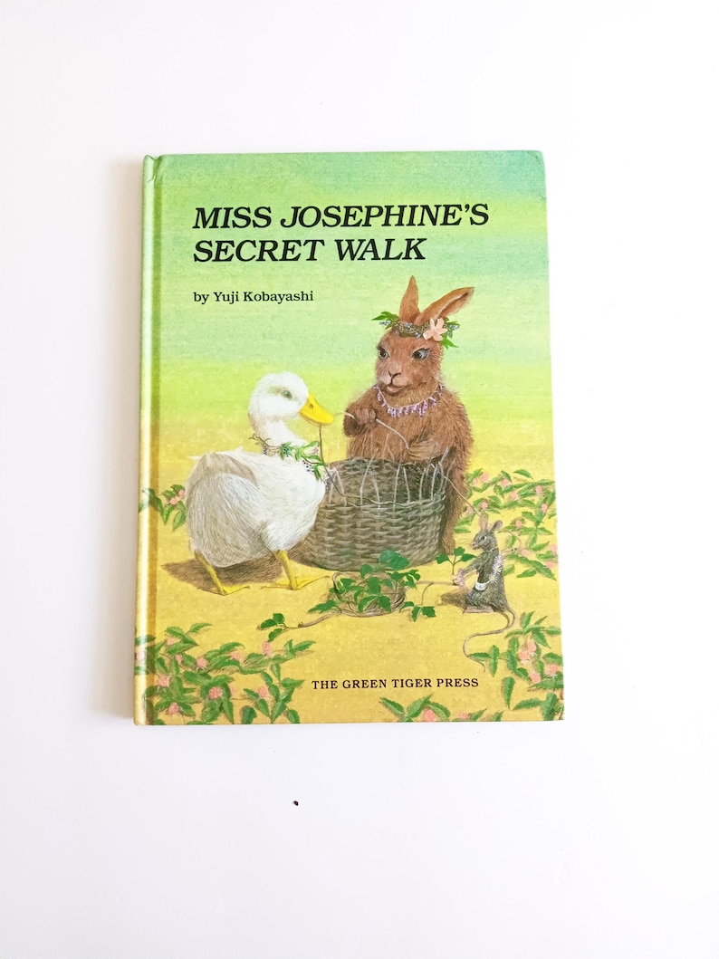 May include: A children's book titled "Miss Josephine's Secret Walk" by Yuji Kobayashi. The cover features a duck, a rabbit, and a mouse near a woven basket. The background is a yellow-green field with pink flowers.