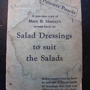 Peut inclure: Une couverture de livre vintage avec le titre "Salad Dressings to suit the Salads". La couverture est imprimée en encre noire sur un fond de papier brun clair. Le texte "Printers Proofs" est estampillé dans un rectangle noir dans le coin supérieur droit de la couverture.