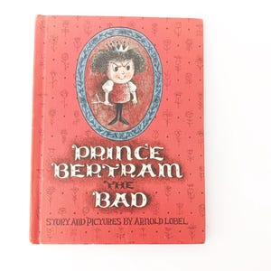May include: A vintage children's book titled "Prince Bertram the Bad" by Arnold Lobel. The cover is red with a floral pattern and features an illustration of a frowning prince in a crown. The book's title is in large, ornate lettering.