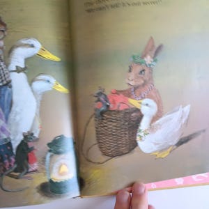 May include: An illustrated page from a children's book featuring anthropomorphic animals. A rabbit, ducks, and a mouse are depicted in a whimsical scene. The rabbit wears a floral crown and necklace, holding a basket. The ducks have yellow beaks.