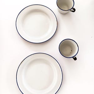 May include: Two white enamel plates and two matching mugs with dark blue rims and black handles are arranged on a white surface. The plates are round, and the mugs are cylindrical.