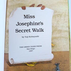 May include: Illustration from a children's book titled "Miss Josephine's Secret Walk" by Yuji Kobayashi. The book cover features a scroll held by brown hands, with a small mouse in the corner. Published by The Green Tiger Press in San Diego, 1987.