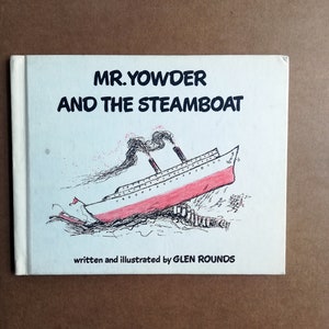 Puede incluir: Una cubierta de libro blanca con texto negro que dice "MR. YOWDER AND THE STEAMBOAT". La cubierta del libro presenta una ilustración en blanco y negro de un barco de vapor rojo y blanco con humo saliendo de la chimenea. El texto "written and illustrated by GLEN ROUNDS" está impreso en negro en la parte inferior de la cubierta.