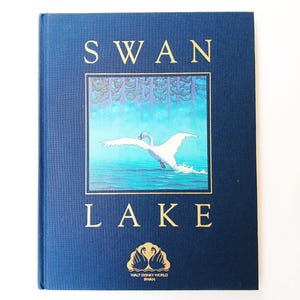 May include: A blue book with the title "SWAN LAKE" in gold lettering. The cover features a painting of a swan flying over a lake with a forest in the background. The book is from Walt Disney World Swan.