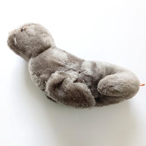 May include: A plush gray seal toy with a soft, furry texture. The toy has a long, slender body, flipper-like appendages, and a small head with a detailed face. The seal is lying down on a white surface.