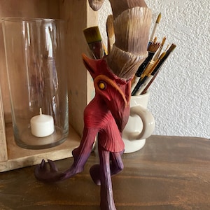 Puede incluir: Una figurita de madera tallada a mano de una criatura con un cuerpo rojo, patas moradas y un cuerno grande y rizado. La figurita tiene un ojo amarillo y está pintada con un degradado de colores.