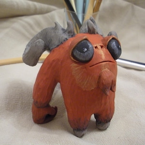May include: A small, red, clay figurine of a monster with horns, a beard, and large eyes. The figurine has a textured surface and is painted with details in brown and gray.