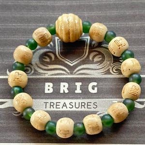 Puede incluir: Una pulsera de cuentas con cuentas de madera de color tostado claro grabadas con caracteres chinos que se alternan con pequeñas cuentas esféricas de color verde oscuro translúcido. Una cuenta de madera más grande y estriada se sitúa arriba.