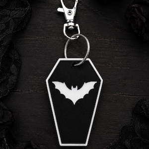 May include: A coffin-shaped keychain featuring a black base with a white border and a white bat silhouette in the center. The charm is attached to a silver-toned metal split ring and a lobster claw clasp for secure attachment to bags or keys.