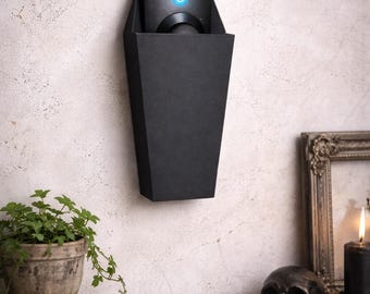 May include: A black, coffin-shaped wall-mounted device with a blue light. The device is mounted on a textured, off-white wall. A small potted plant, books, a skull, and a candle are on a shelf below.
