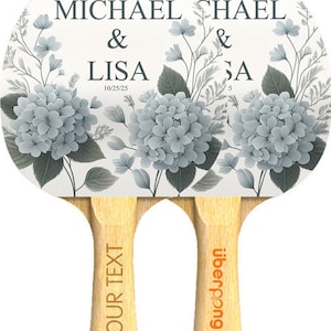 Może przedstawiać: Para białych rakiet do tenisa stołowego z kwiatowym wzorem z niebieskimi hortensjami. Rakietki są spersonalizowane nazwiskami "Michael & Lisa" i "Chael & Sa" oraz datami "10/25/25" i "5". Uchwyty są oznaczone "Your Text" i "Uberpong".